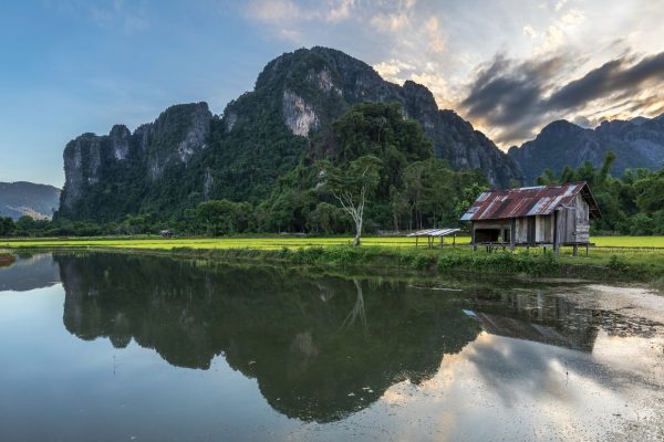 Water_reflection_of_karst_mountains,_wooden_hut,_trees_and_colorful_clouds_at_sunset_with_green_paddy_fields,_Vang_Vieng,_Laos